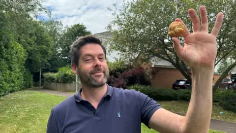 Man holding a scone-like round biscuit in his hand. Looking at the camera. In a small green field. He is wearing a navy top.