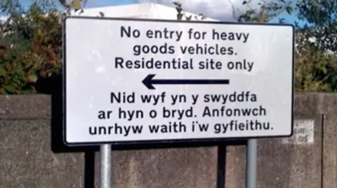 A black and white sign with the words no entry for heavy goods vehicles. Residential site only, with the words "I am not in the office at the moment. Send any work to be translated" in Welsh below
