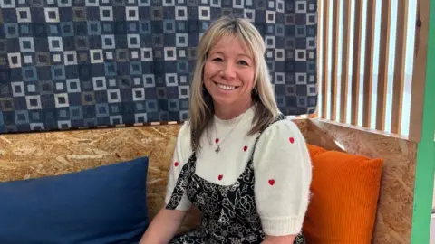 A woman with blond hair sits on a wooden banquette with an orange and a blue cushion. She is smiling at the camera and is wearing a white top with small red hearts.