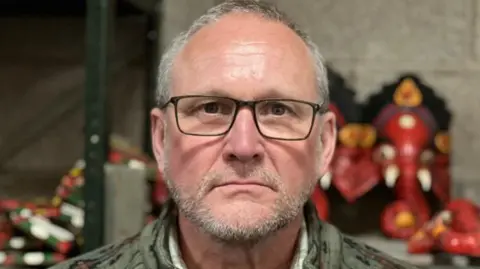 Bernard Rostron. He has short grey hair and beard with black-framed glasses