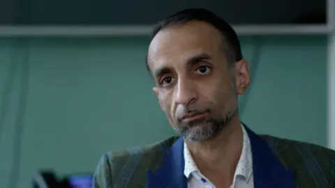 Ben Schofield/BBC Shaz Nawaz looks with an unsmiling expression at the camera. He is wearing a blue-and-green-chequered blazer with a white shirt underneath. He has dark hair, a beard and brown eyes. 