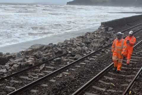 BBC/Johnny Rutherford Engineers in high‑visibility clothing inspecting a section of collapsed sea wall near the Dawlish–Teignmouth railway, with exposed foundations and waves in the background.