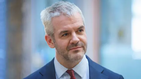 A man with short grey hair and stubble is wearing a blue suit jacket, blue striped shirt and red tie