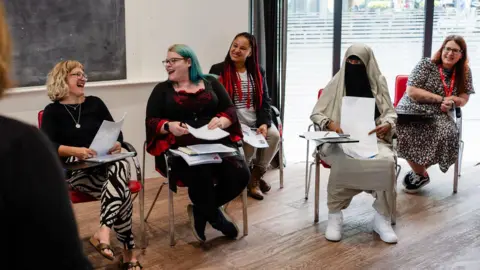 Meg Lovell/Women's Work Lab Five women are sitting in chairs with stacks of paper on their laps or on small desks attached to the arm of the chair. A blonde woman with glasses and a woman with glasses and blue hair are looking at each other and laughing. A woman with long black and red braids is smiling at them. Next to her are a woman with brown/red hair and a woman wearing a Niqab.