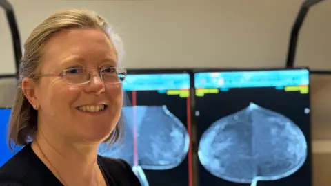 Dr Nerys Forester, a consultant breast radiologist in Newcastle is smiling at the camera. She is wearing glasses and has her blonde hair tied back. Behind her are two screens with soft focus mammogram images. They show how dense tissue can show up as white.