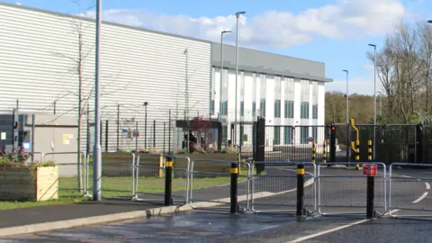 The exterior of a grey and white building with two rows of external temporary fencing and also black and yellow traffic blocking posts