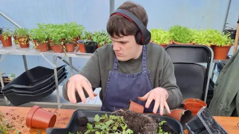 Oakfield Community A man with headphones and a grey sweatshirt and a denim apron tending to some plants