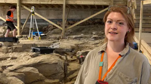 Amy Rattenbury smiling at the camera standing on part of the rocky fossil forest site with a student in the background using equipment to study it. She has long red hair and has glasses perched on top of her head, and wears a brown shirt.