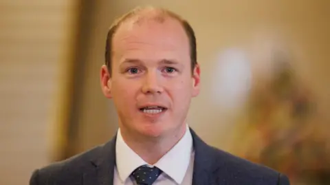 A balding man with blue eyes mid speech with a yellowy blurred background behind him. He is wearing a white shirt, navy tie with pink spots on it and a navy blazer. 