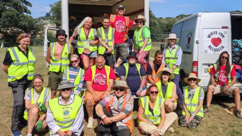 Angus Clark Sixteen men and women wearing high vis jackets either sitting down or standing in front vans which have their rear doors open. One of the vans has a logo which reads "Herts for Refugees". They are in a field with trees and metal fencing in the background.