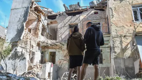 eople stand near a two-story residential building, partially destroyed by a Russian drones' strike on July 12, 2025 in Lviv,