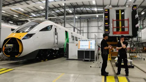 Getty Images Two workers talking inside the Hitachi train factory. A train stands to their right. Its nose is missing and stairs have been placed next to its open doors.