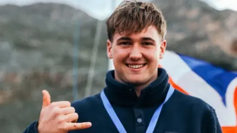 Finn Hawkins has brown hair and is wearing a navy, button up fleece with a medal around his neck. He is holding out his hand with his thumb and pinky finger pointing outwards. He is smiling at the camera.