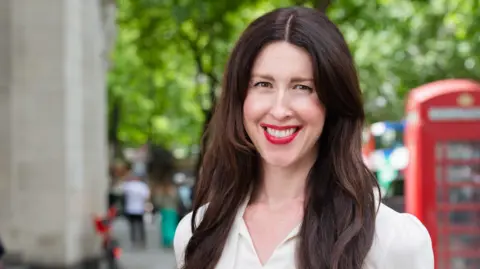 Dr Gillian Brooks A woman with brown hair and a bold red lip is smiling at the camera. There is a typical London background blurred out behind her with a red telephone box, trees, and a concrete building.