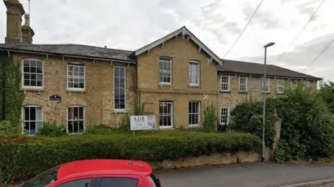 Hill House a yellow Cambridgeshire brick two storey building, possibly originally Victorian. It has white painted sash windows. In front of it is a low wall, covered in greenery and a sign saying ADR. In front of that is a pavement and the glimpse of a red-roofed car