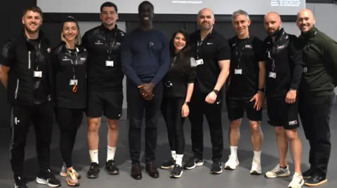 Eastern Education Group Dwain Chambers standing in the middle of a group of men and women, all of whom are looking at the camera smiling. 