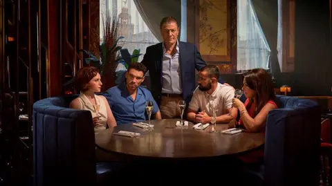 BBC/Left Bank Pictures/James Stack Left to right: Diana Williams (Hannah Onslo), Michael Kavanagh (James Nelson-Joyce), Ronnie Phelan (Sean Bean), Jamie Phelan (Jack McMullen), Elaine Phelan (Julie Graham). They are all seated ina royal blue velvet booth except for Sean Bean who is standing in the centre. The Liver Building can be seen in the background out of the window.