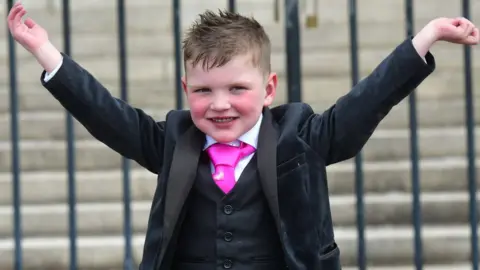 Pacemaker Dáithí MacGabhann smiles with his arms outstretched. He is wearing a black three-piece suit and a pink tie.