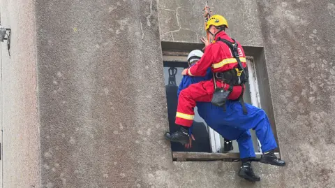 Logan is in a harness infront of a window as he is being lowered down. He is holding onto the casualty. Logan is in a red jumpsuit while the casualty is in a blue one.
