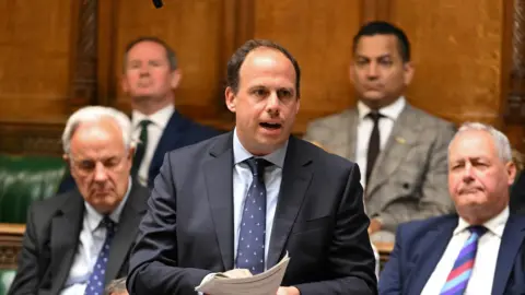 Greg Smith, centre, speaks at the House of Commons in London. There are two other men in suits behind and to each side of him.