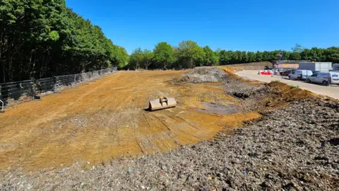 EA More areas of the field flattened and cleared, with rubbish higher at the sides. Vehicles are parked nearby.
