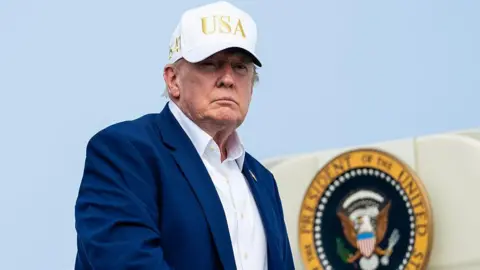 Trump, wearing a suit and no tie and a white "USA" hat, descending from airplane steps