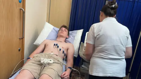 A teenager lies on a bed during a heart scan. There are various wires and electrodes attached to his chest. There is a nurse facing away from the camera next to his bed.