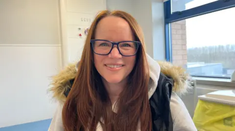 Ellen Knight/BBC Kinder pictured looking into the camera and smiling. She is wearing a light grey hoodie, with a black padded gilet with a light brown fur trim on the hood. She has long, auburn hair, and is wearing a pair of dark-blue-framed glasses. Kinder is sat down in one of the clinic rooms - behind her is a light blue examination bed, and to her left is a yellow and white clinical waste bin. Over her left shoulder is a large, blue-framed window, looking out towards a cluster of trees. The walls of the room are white. 
