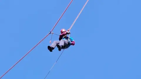 An elderly man abseils down a high building - all that is visible behind him is clear, blue sky. 
