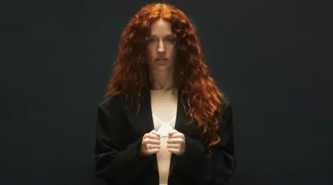 Jess Glynne, with long red hair, is looking at the camera. She is wearing a dark jacket and standing in front of a black background. 