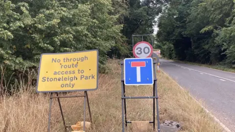 A yellow road sign displaying the words 'No through route access to Stoneleigh Park only' and next to it is blue and red dead end sign. Both are on a grass verge next to a road