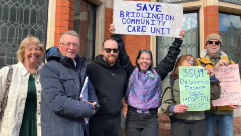 A group of six men and women in outerwear hold up placards with slogans reading: "Save Bridlington Community Care Unit".