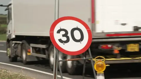 A white lorry drives past a 30mph speed limit sign next to a road.