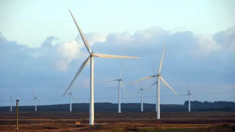 A wind farm on a brown field with white spinning turbines. The sky is cloudy and pale.