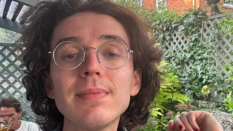 Esme Byrd A picture of a person's face with brown wavy hair and circular glasses with a garden trellis behind and trees climbing up it