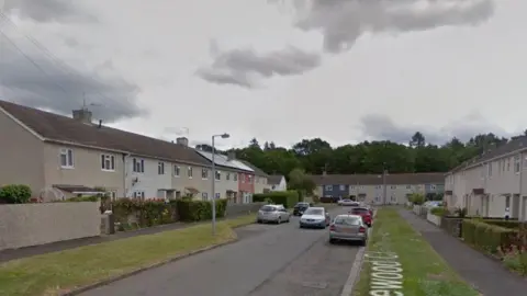 A Google Streetview image of a residential street with rows of terraced houses on the left, right and in the distance. The sky is very grey and there are cars parked along the street. 