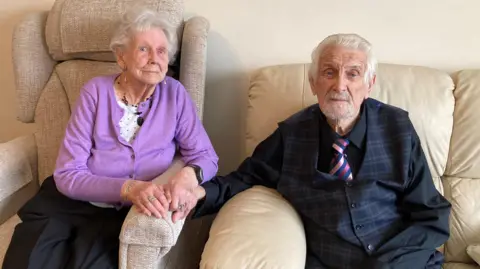 BBC Derek (right) and Brenda (left) Dodge sitting on two chairs side-by-side, holding hands in their home on their 70th wedding anniversary.