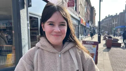 Kate Bradbrook/BBC Lily with long dark hair wearing a light brown fleece top and standing in a shopping street. A Costa A-board is visible behind her and some shoppers are making their way down the street.