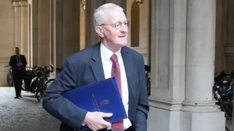 PA Hilary Benn wearing glasses, a navy suit, white shirt and red tie walking carrying a blue folder.