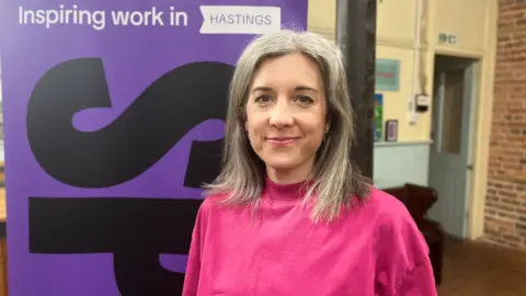 Josh McLaughlin/BBC Ness Moss stands next to a Spear-branded pop-up sign reading: "Inspiring work in Hastings." She smiles for a photograph and is wearing a bright top. She is stood in a room with a brick wall and a light-blue open door.