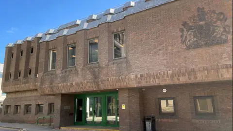 PA Media Chelmsford Crown Court is a brick building with green entrance doors, multiple rectangular windows, and a large royal coat of arms emblem on the wall.