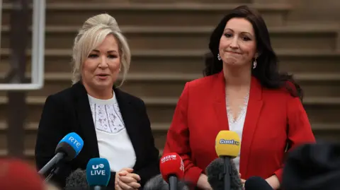 First Minister Michelle O'Neill and Deputy First Minister Emma Little-Pengelly talk to the media. O'Neill has blonde hair, a black jacket and white blouse. Little Pengelly has long dark hair, a red jacket and white blouse.