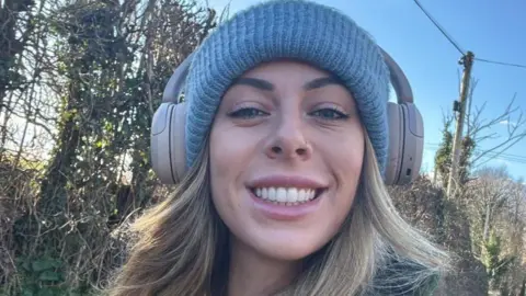 A woman smiles into the camera close up. She has long dark blonde hair under a light blue woolly hat. She is wearing headphones on top of the hat. Behind her are trees, a telegraph pole and blue sky.