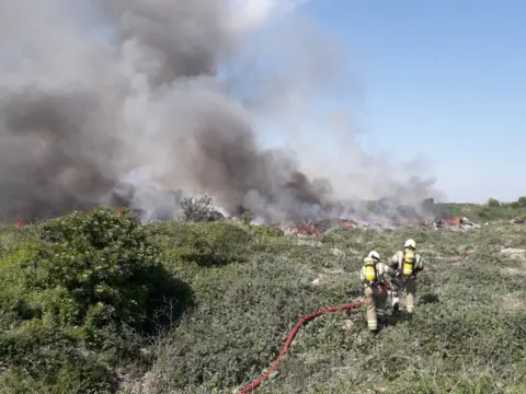 London Fire Brigade Two firefighters use a hose on a fire on scrubland