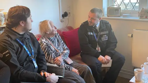 Friends of the Elderly An elderly woman with a black top and wearing a bag over her shoulders sitting on a red sofa with two police officers in uniform sitting either side of her. They are both facing her. There are cups of tea in front of them on a table.