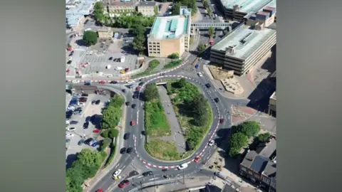 An aerial view of a roundabout with cars driving round it. The roofs of buildings and a car park dot the landscape along with a few with areas of trees and grass. 