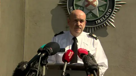 A police officer is standing in front of a podium speaking to the media. Behind him, the PSNI emblem is on a wall. A number of mics are in front of him.