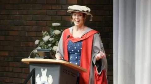 University of York Jones wears a grey and red graduation gown with a large grey cap. She stands at a grey lectern and speaks into a microphone. behind her is a brick wall with white flowers in front of it.