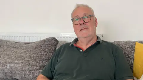 A man sat down on a cream sofa, wearing a green polo short and cream trousers, clutching his mobile phone.