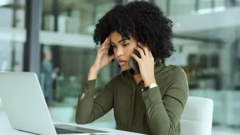 Getty Images A stock image of a woman holding a mobile phone to her ear while looking at her laptop. She looks concerned.